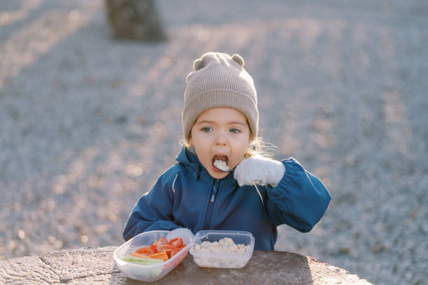 istock Little girl eats porridge with a spoon while standing behind a large tree stump with a lunchbox 2154330849