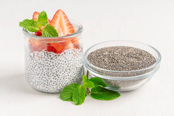 Glass jar with chia pudding dessert decorated with strawberries and mint leaf and bowl with seeds on white table Sweet healthy chia pudding dessert made with milk decorated with topping of fresh mint leaves and sliced ripe juicy strawberries served in glass jar with bowl of seeds ingredient on white wooden table eating chia seeds stock pictures, royalty-free photos & images