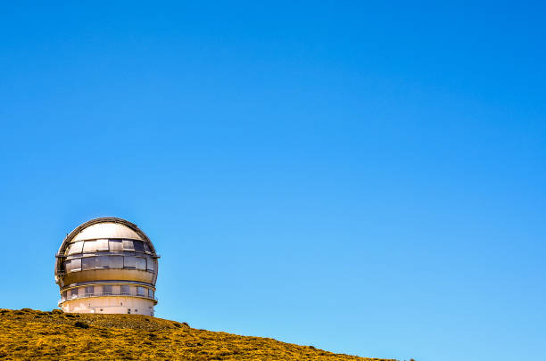 A large telescope is on a hill with a clear blue sky. The telescope is old and has a weathered appearance. The scene is peaceful and serene, with the telescope standing alone on the hilltop