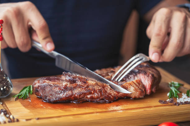 Men's hands holding knife and fork, cutting grilled steak Hands holding fork and knife and eating delicious juicy steak on a wooden plate steak stock pictures, royalty-free photos & images