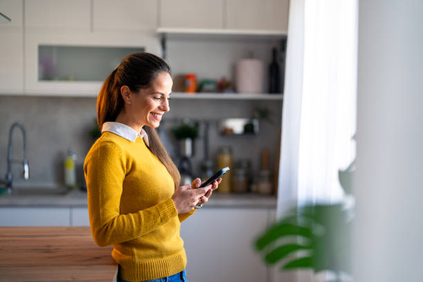 istock Smiling young businesswoman sending messages over smart phone while standing at kitchen counter 2154358339