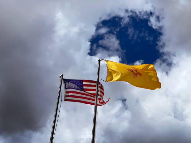 The United States flag and the New Mexico state flag, mounted on flagpoles, fly in the wind under a stormy, cloudy sky, but with blue sky opening up through the clouds above the state flag. The brightening sky allows sunlight to hit the surfaces of the flags and enhance their rippling folds. The stars and stripes of the national flag, with its red, white, and blue colors, complement the bright yellow background and red graphic stylized sun on the state flag. The state sun symbol is reminiscent of Native American hieroglyphic drawings of the sun.