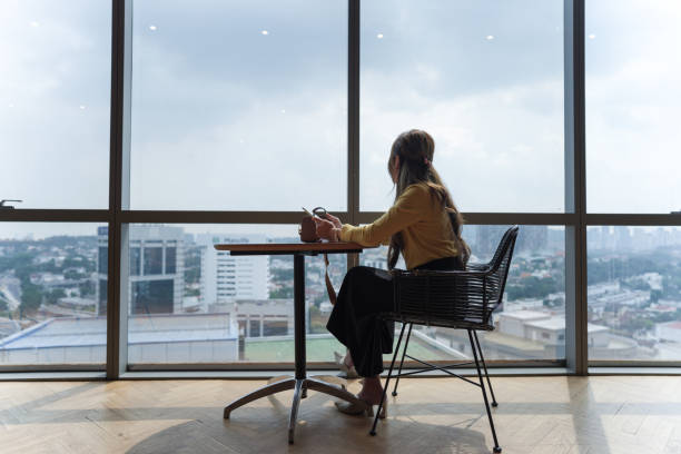 istock A woman in a yellow sweater sits at a table in front of a large window using a digital tablet with a cityscape in the background 2154371855