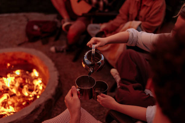 Close up shot of a woman pouring a man some tea from a shiny, metal kettle while sitting near a campfire.