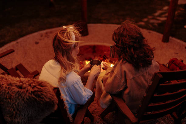 Brunette and a blonde share a moment drinking coffee by a fire pit.