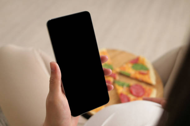 istock Unrecognizable pregnant female eating pizza and using mobile phone holding smartphone with black blank screen advertisement of delivery service of fast food 2154380456