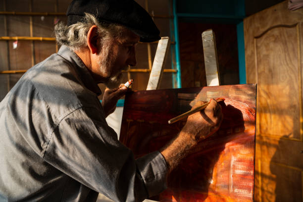 Older man between 55-60 enters his closed-door studio to create art and make a painting on canvas, while the afternoon falls and the shadows of the sun are seen on the window that gives a nostalgic atmosphere to his creation