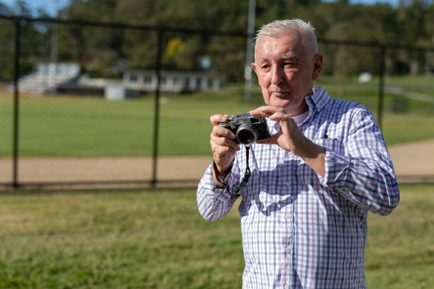 Elderly male photographer using a digital DSLR camera at eye level. Blue shirt against a soft background of green trees