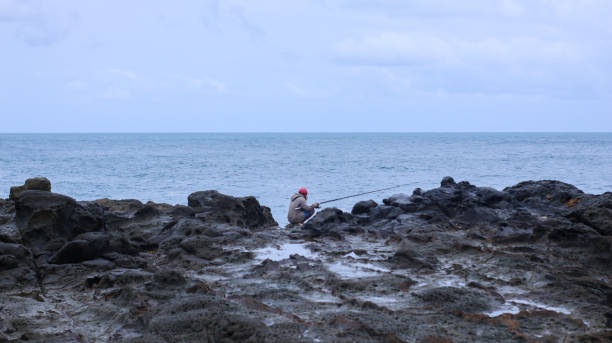 Yogyakarta, Indonesia - September 20, 2022 : A solitary man stands fishing on the weathered rocks by the sea
