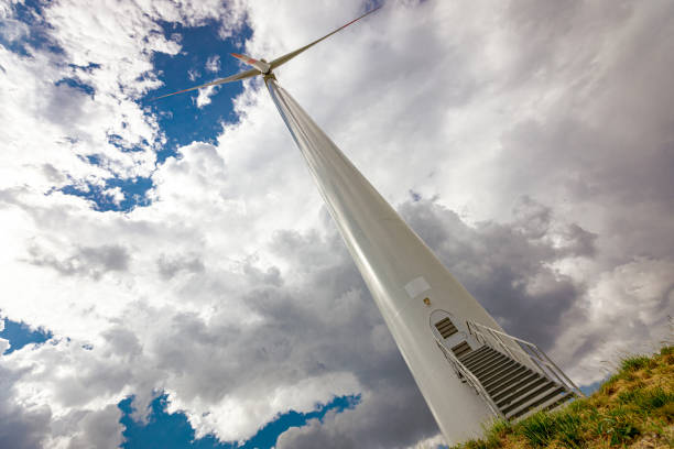 Front view on entrance, doorway with stairs into large wind power turbine with rotating blades, propellers, against cloudy stormy sky. Producing renewable clean electricity converting kinetic energy.
