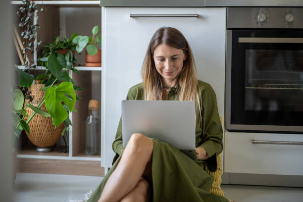 istock Pleased relaxed woman smiling using laptop on knees sitting on kitchen floor chatting, scrolling web 2154417804