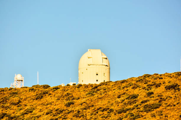 A large white telescope is on a hill with a clear blue sky. The telescope is the main focus of the image, and the hill and sky provide a serene and peaceful backdrop