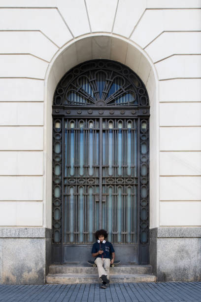 Anonymous male with legs crossed relaxing on skateboard and looking at screen of mobile phone while sitting on steps of building huge arched entrance with grill door on street in daylight