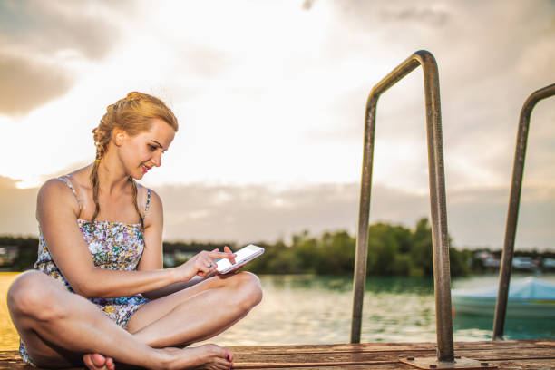istock Young woman using digital tablet while sitting by the lake at sunset 2154439401