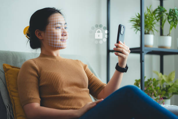 kuvapankkikuvat ja rojaltivapaat kuvat aiheesta asian woman scanning face id by smart phone sitting on sofa in living room at home. - verifikaatio
