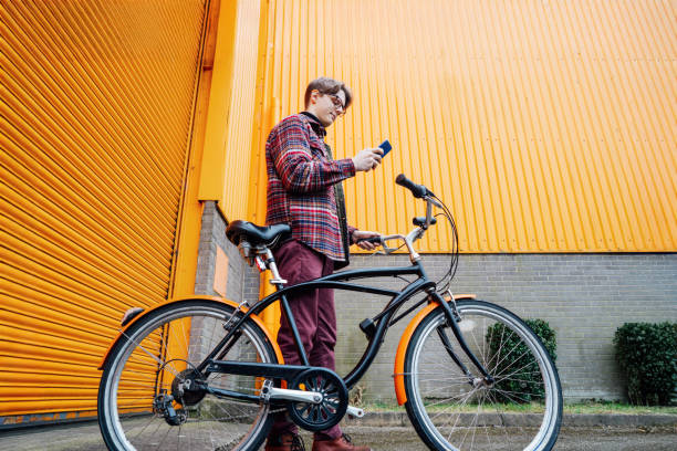 istock Young hipster man using mobile phone while standing near bicycle near orange wall. Student using 5g network,rests after riding. Eco friendly transport for carbon footprint. Sustainable lifestyle. 2154472807