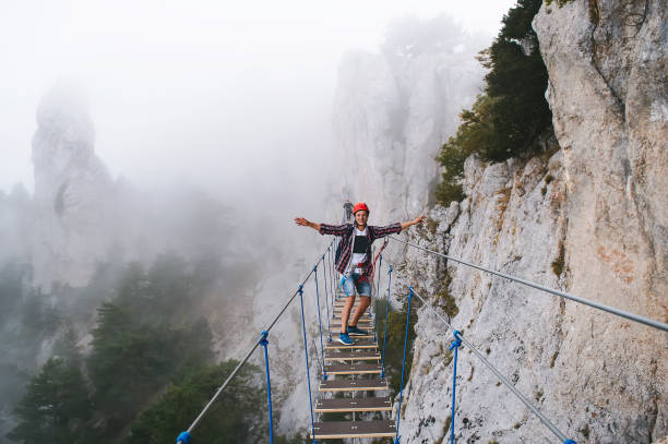 A young man in mountaineering equipment walks uphill on a suspension bridge in the fog. VIA Feratta - Heavenly trail. Suspension bridge on Mount Ai-Petri, Yalta, Crimea. Sea coast in Crimea. The concept of travel, active lifestyle. un joven con equipo de montañismo camina cuesta arriba por un puente colgante en medio de la niebla. via feratta - sendero celestial. puente colgante en el monte ai-petri, yalta, crimea. la costa del mar en la crimea. el concepto de viaje, estilo de vida