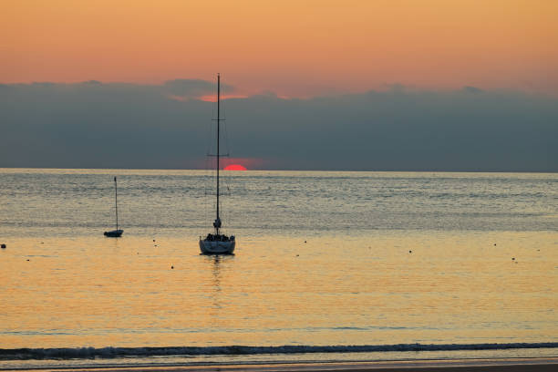 Sailboats at sunset with dramatic sky and red sun rolls over the horizon