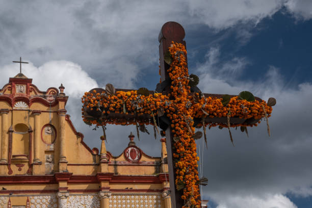 This evocative image captures the essence of the Day of the Dead celebrations in Chiapas, Mexico. In the lower left corner, the upper edge of the San Cristobal Cathedral peeks into the frame, its historic architecture a testament to the region’s rich cultural heritage. Dominating the center of the image, without touching the cathedral, is a part of the atrial cross, adorned with vibrant marigold flowers - the traditional ‘Flor de Cempasúchil’. This poignant symbol of life and death, set against the backdrop of the cathedral, creates a powerful visual narrative of this deeply revered Mexican tradition.