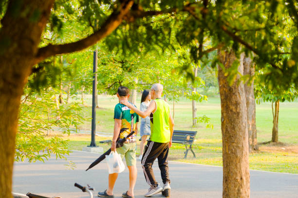 Two thai senior men are walking close together in park Wachira Benchathat Park (Rot Fai Park) in Bangkok Chatuchak. Both are wearing casul and sports clothing. One man is wearing sports shoes. He has arm around sholder of other man. Other man is carrying a small bag and an umbrella. In background are trees and a young woman is passing by in background