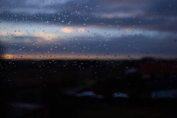Full frame shot of rain drops on window with defocused landscape and cloudy sky in background at dusk seen through glass