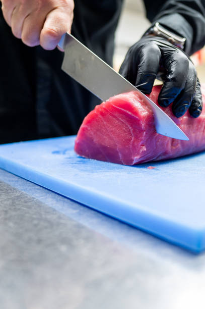 Close-up of hands wearing black gloves slicing red tuna fish on a blue cutting board with a metal knife, depicting food preparation