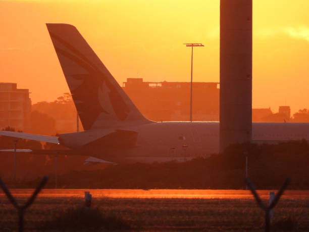 A Qatar Airways Airbus A380-861 plane, registration A7-APL, taxiing after landing at Sydney Kingsford-Smith Airport as flight QR908 from Doha. In the image is a barbed wire fence, the column of the airport's weather measuring installation and residential buildings.  This image was taken from Mill Stream Lookout, Botany Bay with a blood orange coloured sunset on 25 May 2024.