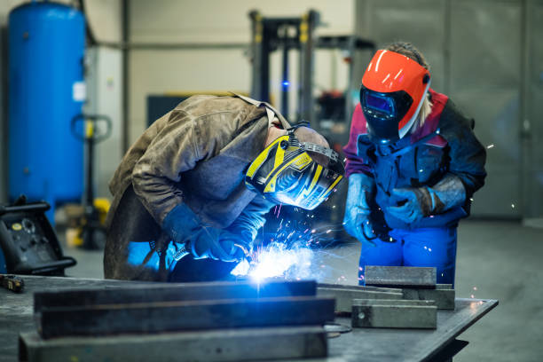 Blonde woman works as a welder in workshop, operating welding machine, wearing protective clothing and a welding mask.