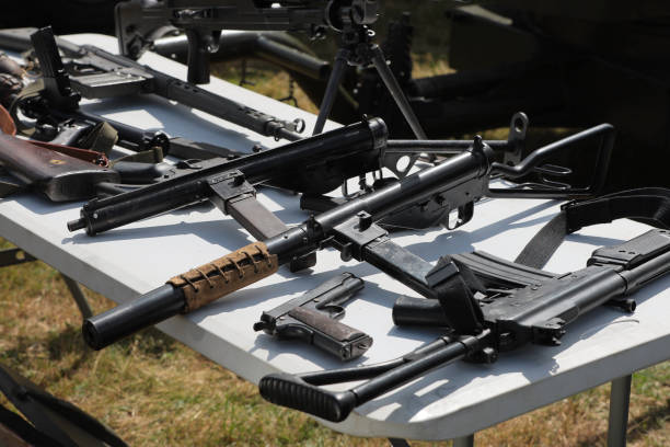 A black machine gun with a long barrel is on a grassy field. The gun is surrounded by a lot of ammunition
