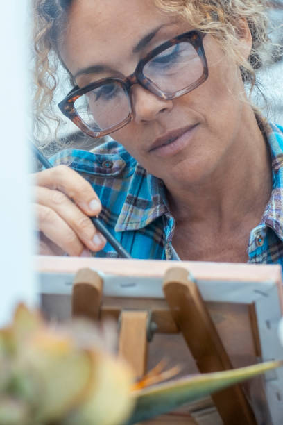 istock Concept of art, creativity, hobbies, work and creative occupation. Front view of blonde, curly woman sitting in front of easel, painting with brush and colors 2154620540