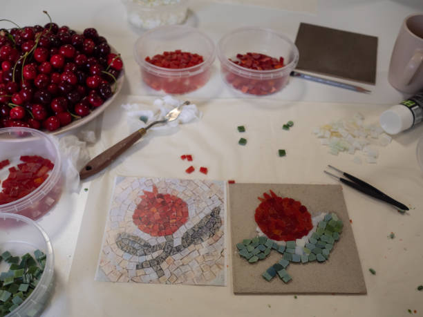 istock Mosaic tools, mosaic base, an out print of mosaic pattern, containers with red, green and white glass pieces, and a bowl of ripe cherries on a white surface. 2154632076