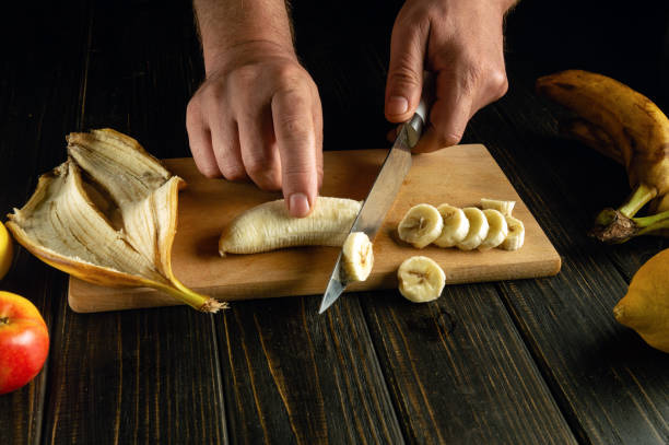 istock Preparing a fruit dish on the kitchen table. Chef hands using a knife to slice a banana on a wooden cutting board 2154641493