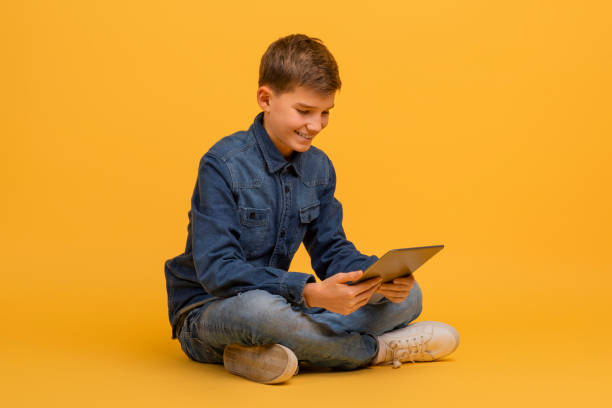 istock Teen boy using digital tablet while sitting cross-legged on floor in studio 2154643451