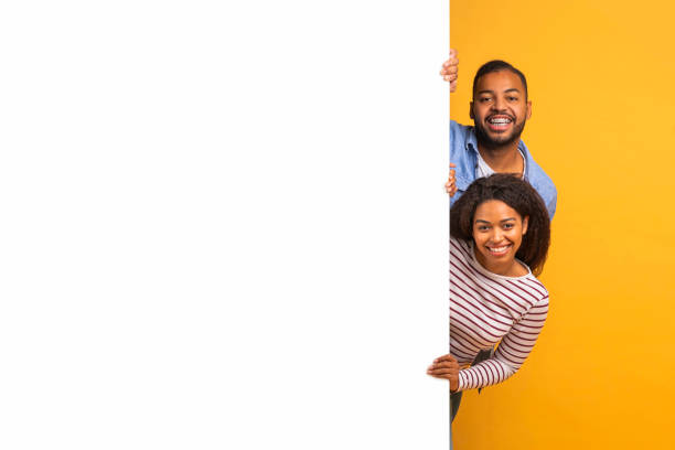 istock Interesting Offer. Happy Young Black Couple Peeping Out Behind White Advertisement Board 2154645079