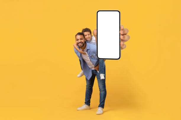 Smiling Black Father And Preteen Son Demonstrating Smartphone With Blank White Screen Smiling Young Black Father And Preteen Son Demonstrating Smartphone With Blank White Screen At Camera, Happy Dad And Preteen Male Child Embracing And Recommending New Mobile App, Copy Space, Mockup kid holding fathers hand stock pictures, royalty-free photos & images