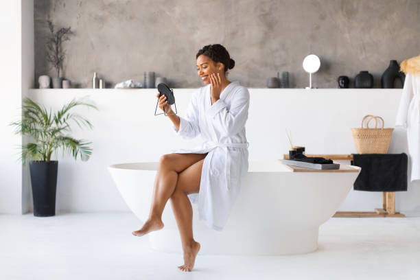 istock Attractive young black woman sitting on bathtub and looking into handheld mirror 2154649544