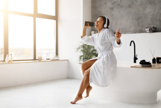 istock Black woman in white bathrobe singing into her phone as microphone, 2154649551