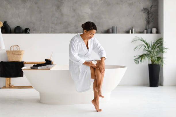istock Smiling Black Woman In Bathrobe Moisturizing Legs With Moisturizer Cream In Bathroom 2154649563