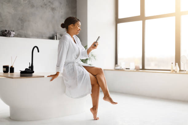 istock Black woman in white bathrobe laughing while texting on her smartphone 2154649568