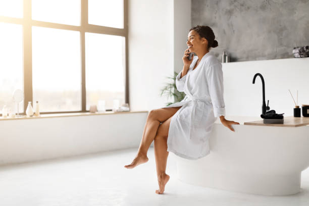 istock Black woman in bathrobe laughing joyfully while talking on cellphone in bathroom 2154649581