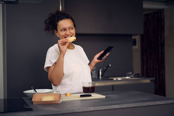 istock Beautiful multi ethnic woman 40s, using smartphone, checking social media content, browsing web, scrolling newsfeed, standing at kitchen counter with a sandwich in hand, enjoying her breakfast at home 2154658672