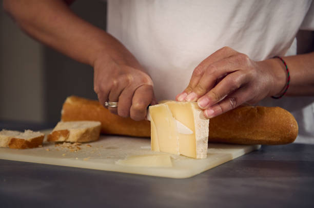 Close-up hands of a woman slicing cheese on cutting board, using a kitchen knife, preparing sandwiches with a loaf of whole grain wholesome integral bread baguette
