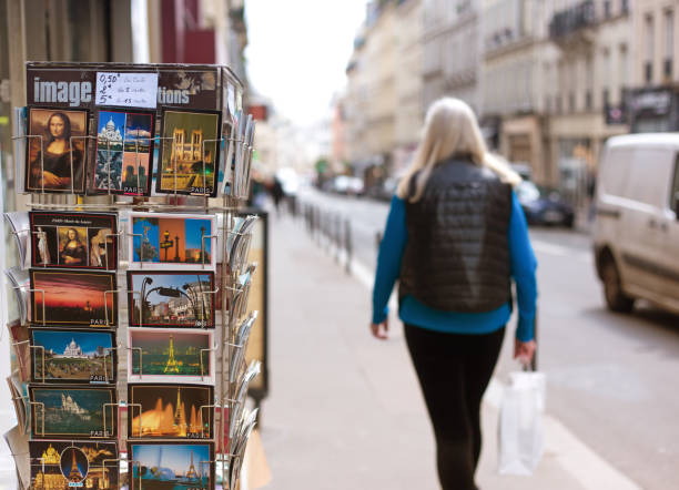 Paris, France: A woman walks past a postcard display stand in the 9th arrondissement showing postcards of Paris icons, including the Mona Lisa, the Eiffel Tower, Notre Damed de Paris, Sacre Coeur, and others.