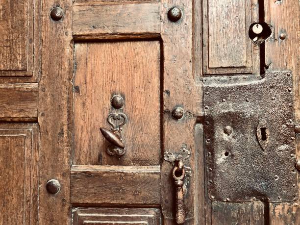 Horizontal closeup photo of part of an old brown timber front door with a nail studded metallic keyhole plate, an ancient doorbell, latch and a modern keyhole. Saumur, Loire Valley, France.