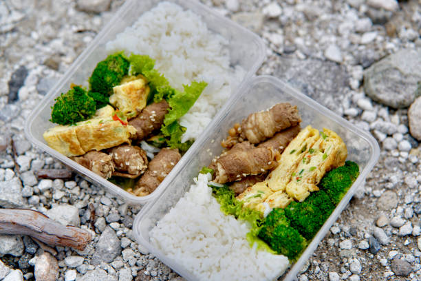 Delicious beef enoki roll, omelette, steamed brocolli, and rice in plastic lunch box. In the background, a white-yellow-sulphuric gravel ground was shown.