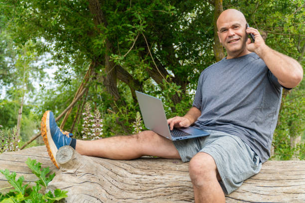 istock A smiling man sitting on the trunk of a tree, using the computer and the phone 2154687291