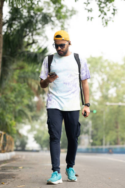 A young man walks along a city street, engrossed in his smartphone. He is dressed in a casual yet stylish outfit, featuring a tie-dye T-shirt, navy cargo pants, and turquoise athletic shoes. He accessorizes with a yellow baseball cap worn backward, sunglasses, and a black backpack. The man holds another small device in his left hand, embodying a tech-savvy urban lifestyle. The background includes lush greenery and a blurred cityscape, indicating an outdoor urban setting