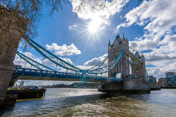 Panoramic view of the River Thames with blue sky and the sun with its rays coming out from behind the clouds backlighting London's Tower Bridge with calm water.