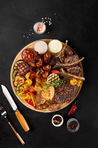 istock Sumptuous BBQ Meat and Vegetable Assortment on Grill Board with Condiments on Black Background 2154700401