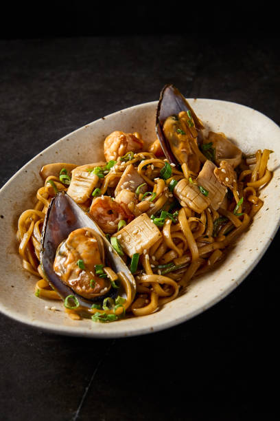 istock A close-up, vertical side shot emphasizing the rich textures of udon pasta with seafood, curated in Asian tradition, on a ceramic plate over a minimalist black tableau 2154701917